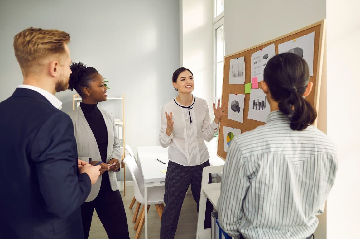 Cuatro personas de un equipo conversan de pie frente a un tablero de corcho con gráficos impresos.