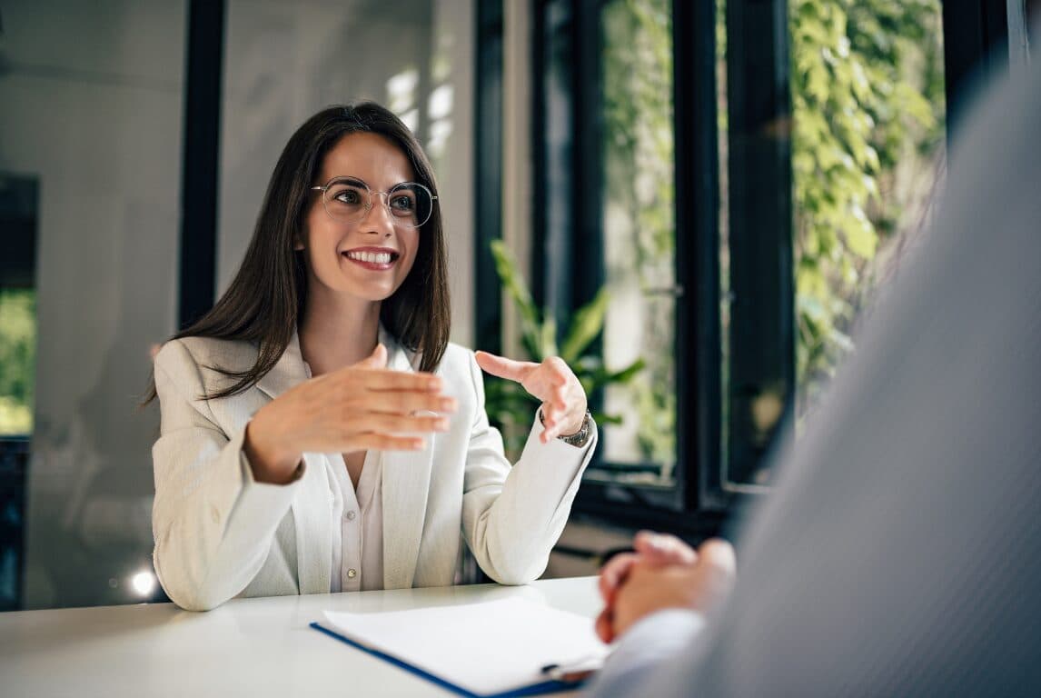Mujer con lentes conversa sonriendo y gesticula con las manos en una sala luminosa.