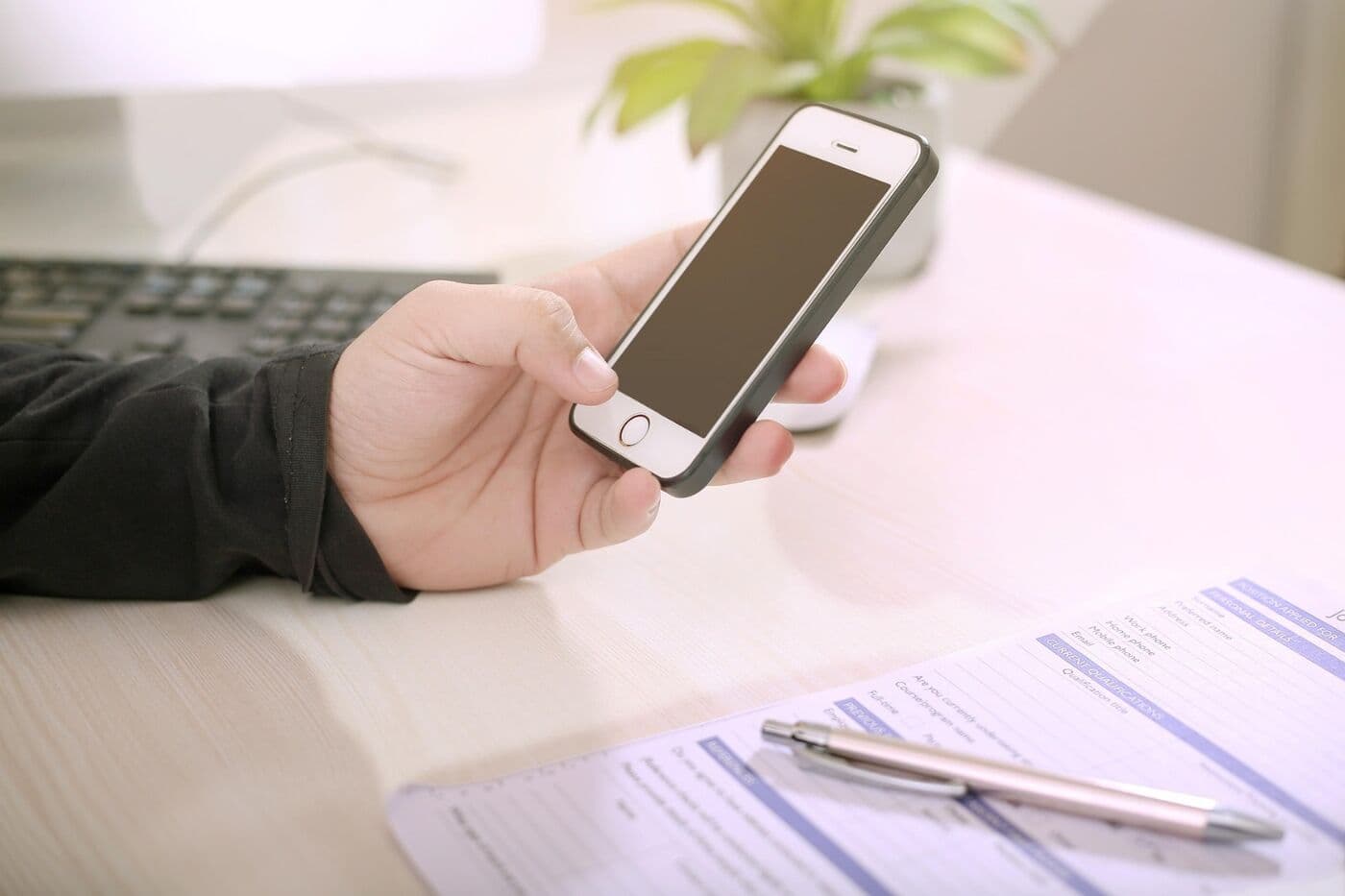 Mano sostiene un teléfono móvil sobre un escritorio, junto a un formulario impreso y un bolígrafo.